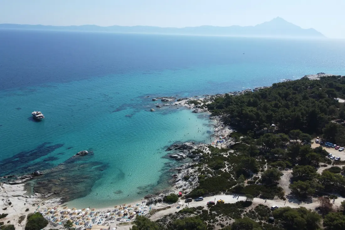 Luftaufnahme von Kavourotrypes: helle Felsen, kleine Buchten, dicht an dicht stehende Sonnenschirme und ein Parkplatz am Hang, am Horizont der Berg Athos